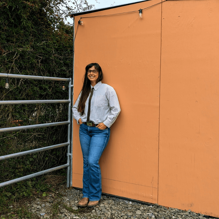 Photo of author, a woman wearing jeans, brown and white striped button up shirt, and a skinny tie, leaning on the horse shelter she and her husband built themselves.
