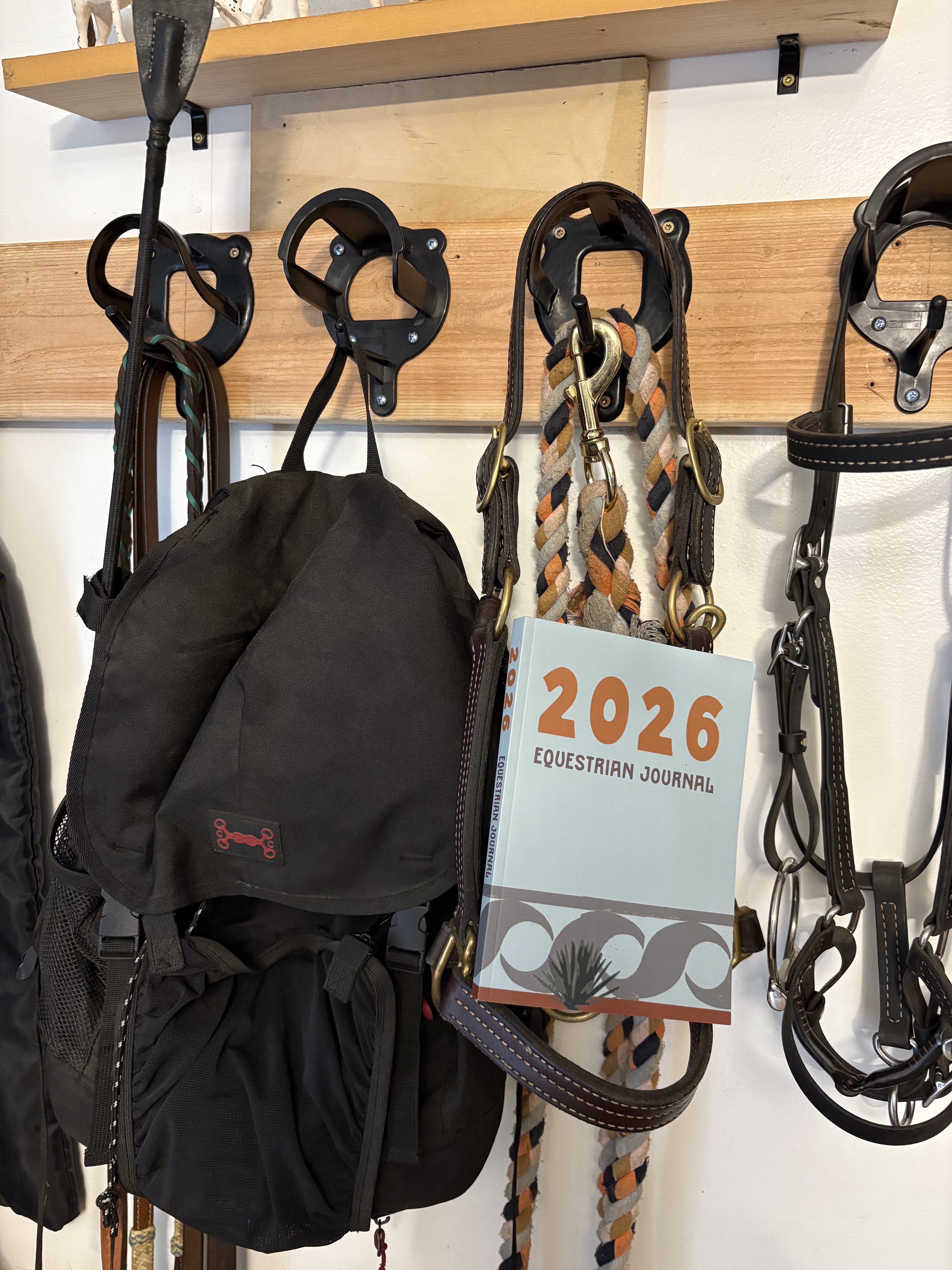 Photo of 2026 Equestrian Journal propped on a leather halter hanging next to a backpack in the tack room.