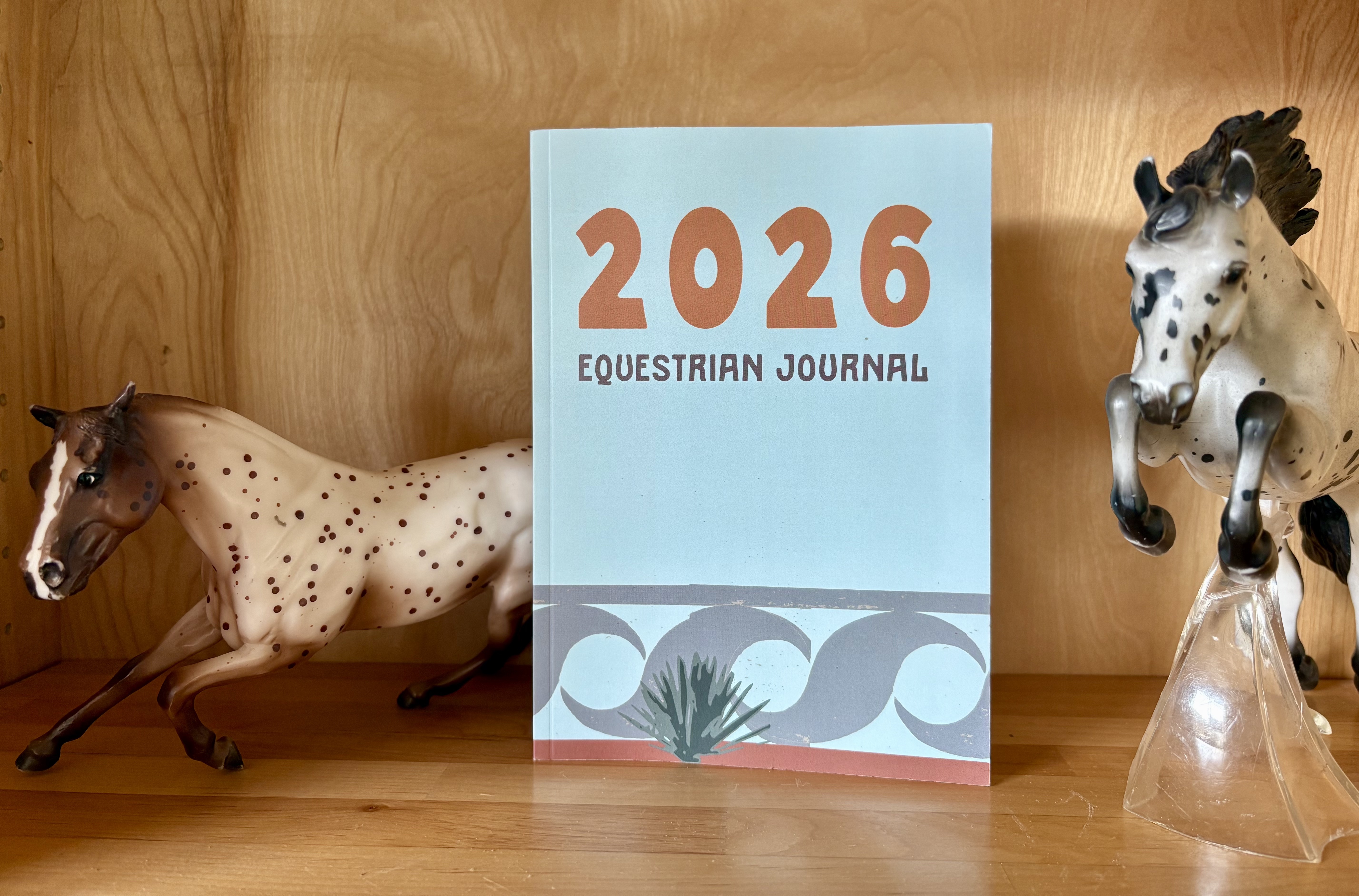 Photo of the 2026 Equestrian Journal on a bookcase between a barrel racing horse figurine and a showjumping horse figurine.