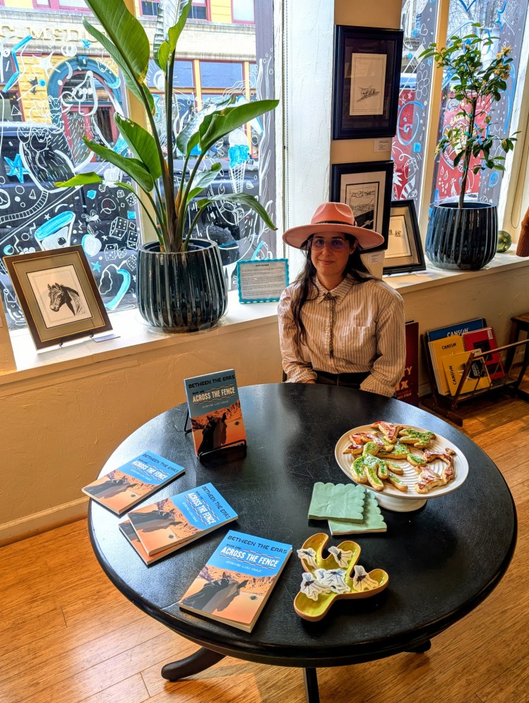 Photo of Stevie Lou Díaz sitting at a table in front of her illustrations at the book release of Across the Fence. 